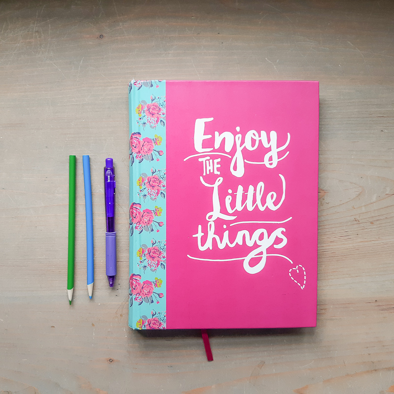 on a wood neutral table, a notebook that says "Enjoy the little things" with a gel pen and 2 color pencils