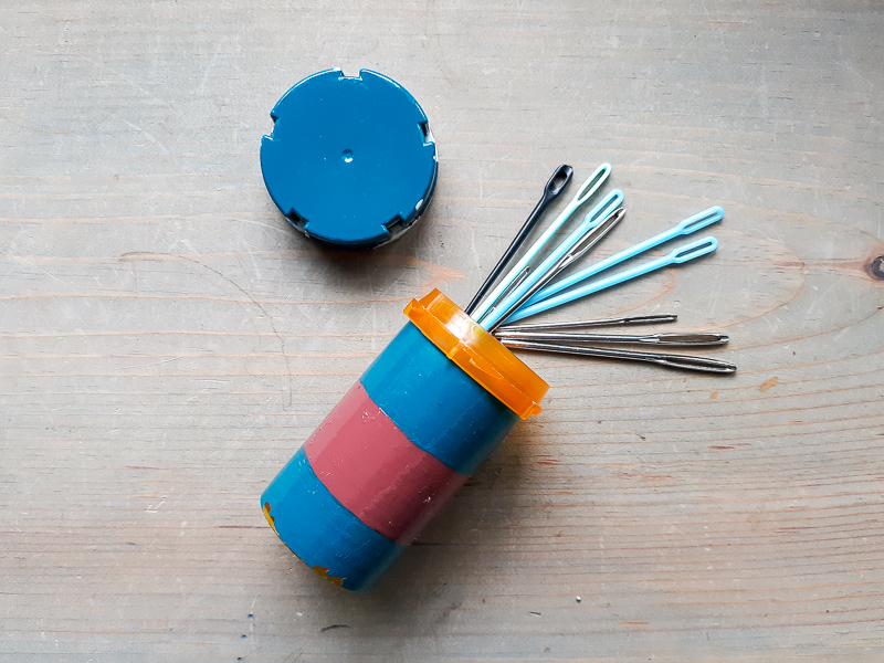 Metal and Plastic Darning needles spilling out of a painted prescription bottle on a neutral wood background